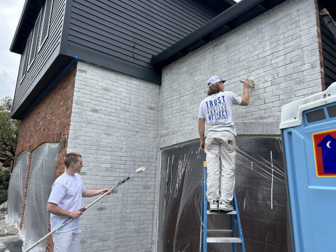 Two painters from Pearl Painters paint a house exterior white in Portland; one on a ladder, one on ground. Portable toilet shown.