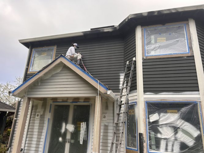 Painter in protective gear spray painting a two-story house in Portland; Pearl Painters sign visible, windows covered with plastic.