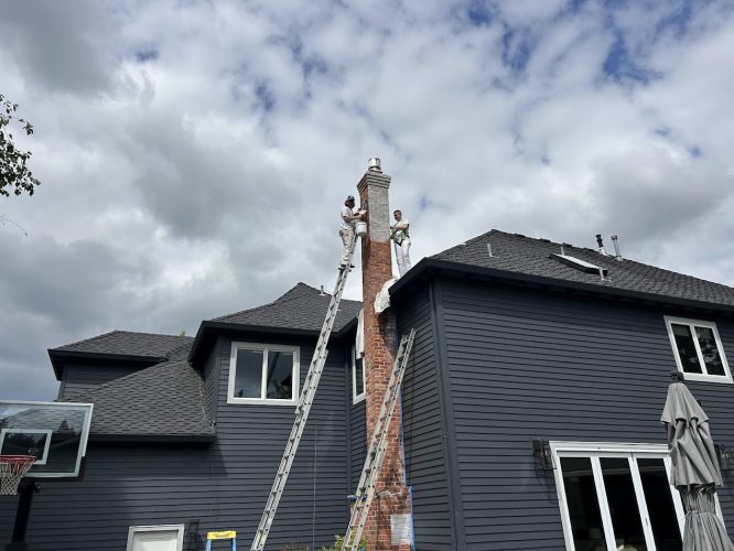 Two painters on ladders work on a brick chimney atop a dark gray house under cloudy skies, likely in Portland. Pearl Painters.