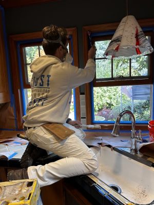 A painter kneels on a kitchen counter in Portland, painting window trim above a sink. Pearl Painters safety gear is worn.
