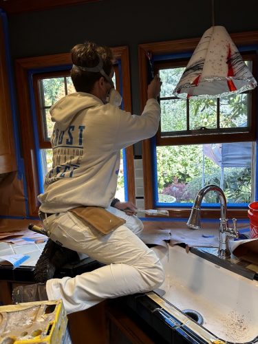 A painter kneels on a kitchen counter in Portland, painting window trim above a sink. Pearl Painters safety gear is worn.