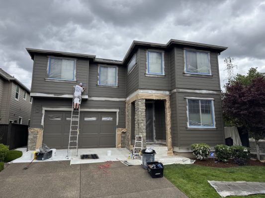 A painter on a ladder paints a two-story brown house in Portland, with windows and garage masked. Painting tools and ladders from Pearl Painters are on the driveway.