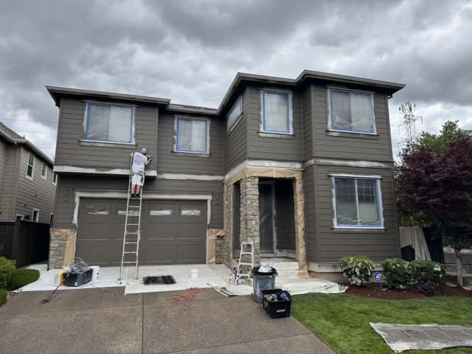 A painter on a ladder paints a two-story brown house in Portland, with windows and garage masked. Painting tools and ladders from Pearl Painters are on the driveway.