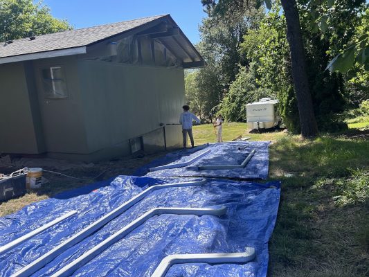 Two painters from Pearl Painters stand near a house in Portland with blue tarps on the grass holding long metal pipes; an RV is parked nearby under trees.