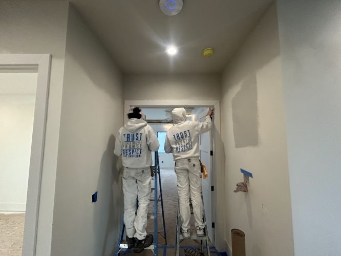 Two painters from Pearl Painters in Portland paint a hallway wall gray, with blue tape on edges and supplies on the floor.