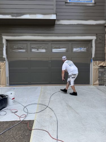 A painter prepares to paint a taped-off garage door in Portland, with coverings and supplies on the ground. Pearl Painters branding not shown.