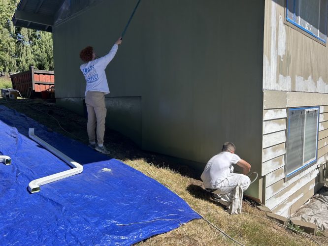 Two painters are painting a house exterior; one rolls paint high up, the other works near the ground. A blue tarp is on the grass. The scene is in Portland with Pearl Painters.