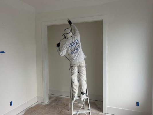 A painter with a spray gun paints doorway trim in an empty, unfinished room. Job by Pearl Painters in Portland.