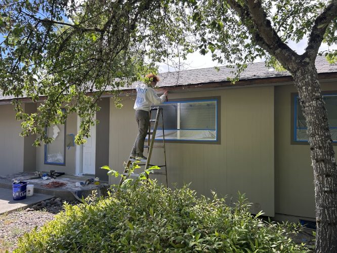 A painter stands on a ladder painting beige house trim, with blue tape on windows and door. Shrubs and a tree are in front. Pearl Painters, Portland.