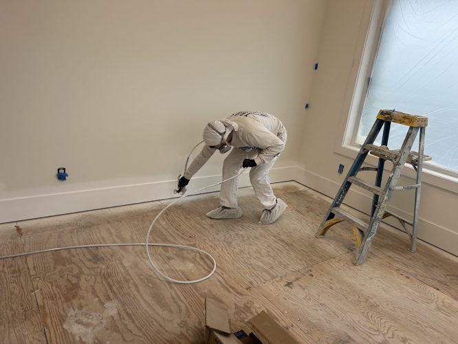 A painter in protective gear spray paints baseboards in an unfinished room; ladder by a window. Pearl Painters, Portland.