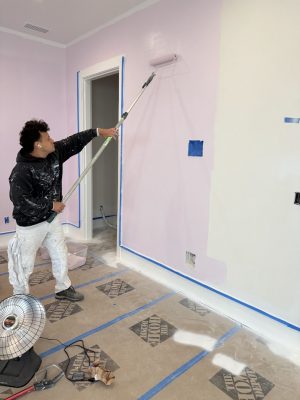 Painter rolls light pink paint on wall; protective sheets cover floor, blue painter's tape lines edges. Pearl Painters, Portland.