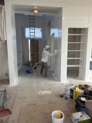 A painter in protective gear sprays white paint on cabinetry amid scattered tools; a Pearl Painters project in Portland.