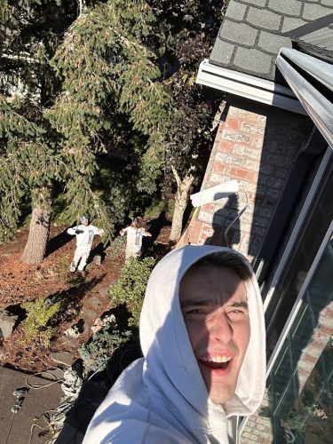 A man in a white hoodie takes a selfie on a high ledge as two painters in coveralls from Pearl Painters are below, painting a house in Portland.