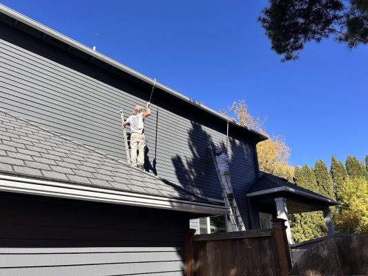 Two painters from Pearl Painters paint a gray house exterior in Portland, one on the roof and one on a ladder, under blue sky.