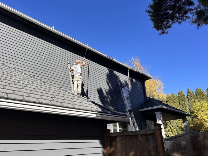 Two painters from Pearl Painters paint a gray house exterior in Portland, one on the roof and one on a ladder, under blue sky.