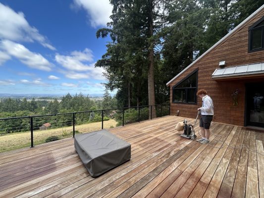 A painter uses a floor sander on a wooden deck next to a modern house, trees and scenic landscape in the background; Pearl Painters, Portland.
