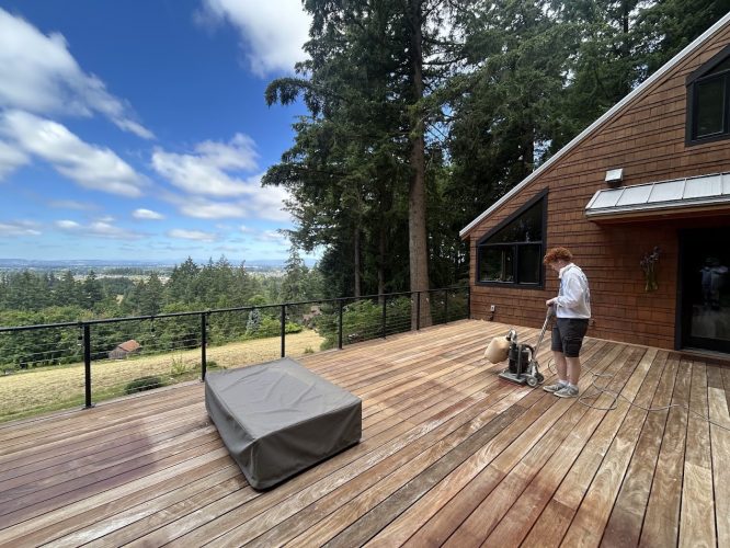 A painter uses a floor sander on a wooden deck next to a modern house, trees and scenic landscape in the background; Pearl Painters, Portland.