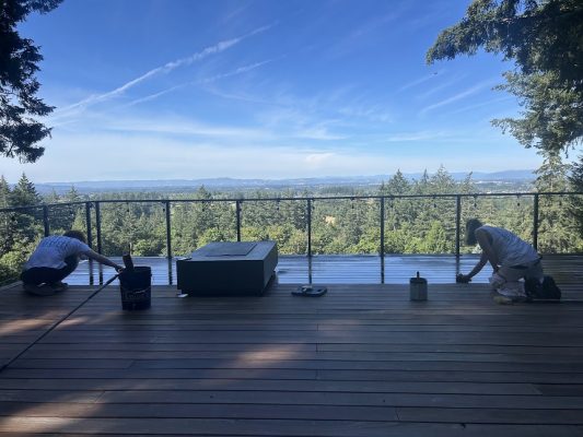 Two painters from Pearl Painters are painting a wooden deck with a railing, overlooking trees and hills under a blue sky near Portland.