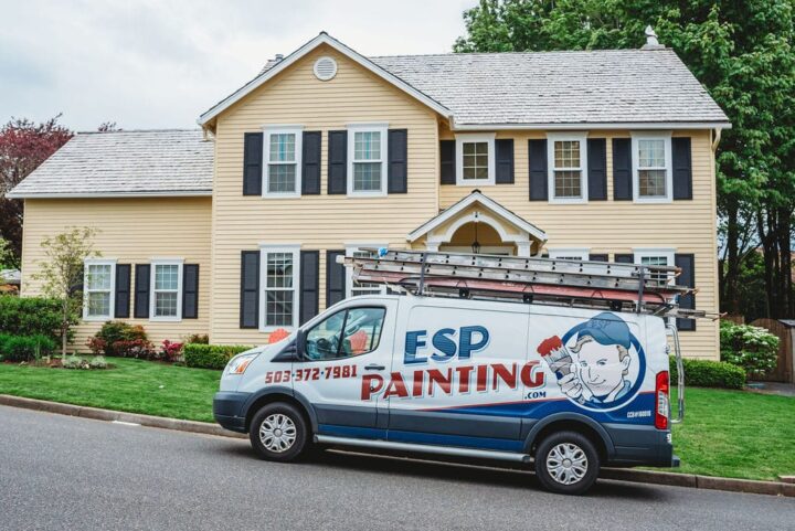 A yellow house with black shutters stands in the background. In front, a white van labeled "ESP Painting" is parked, ladders on its roof. Local Portland painters are on the job, adding fresh touches to the charming home.