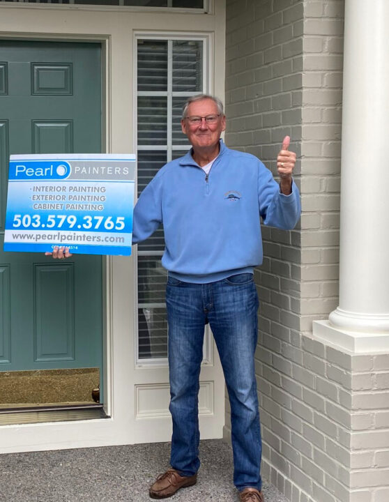 Man with Pearl Painters sign giving thumbs up in front of door; sign lists Portland painting services and phone number.