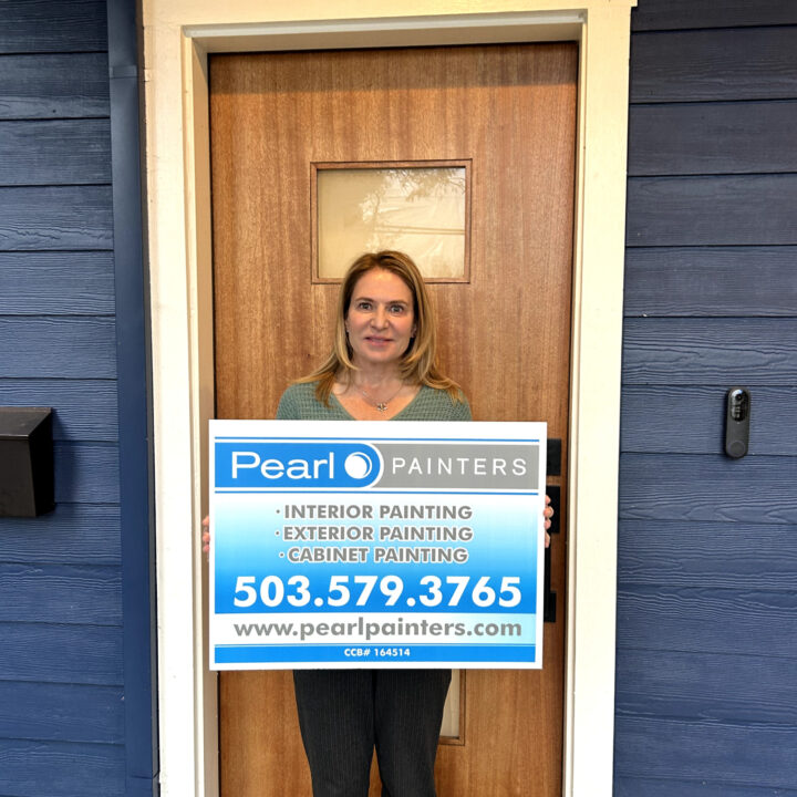 Woman in front of wooden door holds Pearl Painters sign with painting services and contacts; location appears to be Portland.