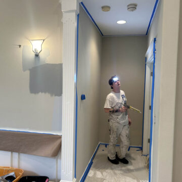 A person painting a hallway wall blue, with painter's tape and a drop cloth in place, under artificial lighting.