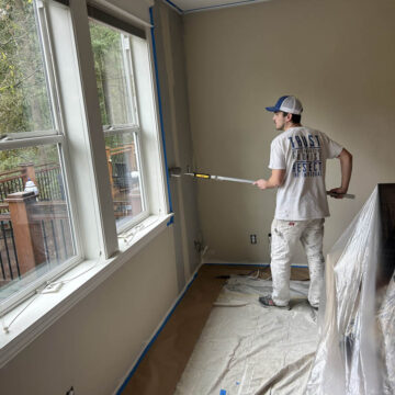A painter, wearing a cap and white overalls, applies paint on a wall in a room with large windows overlooking a wooded area.
