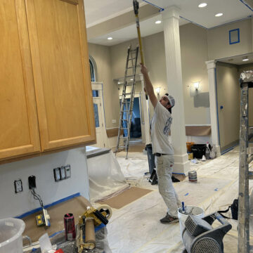 A painter in white overalls using a roller on an extended pole to paint a ceiling in a room covered with protective sheets and painter's tape.