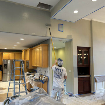 A person in protective gear painting a ceiling near a ladder in a kitchen undergoing renovation.