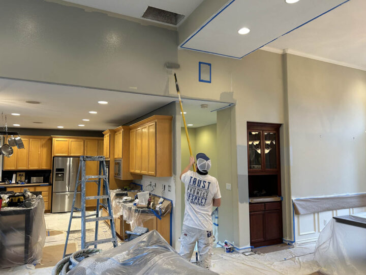 A person in protective gear painting a ceiling near a ladder in a kitchen undergoing renovation.