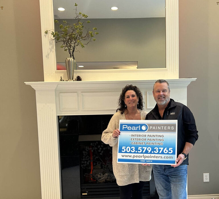 Two individuals posing with a sign for pearl painters, standing in front of a fireplace, showcasing their latest interior painting project in Tigard that brightened the space.