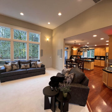 Spacious living room with a large gray sectional sofa, hardwood floors, and adjacent open kitchen with warm lighting. large windows offer a view of greenery outside.