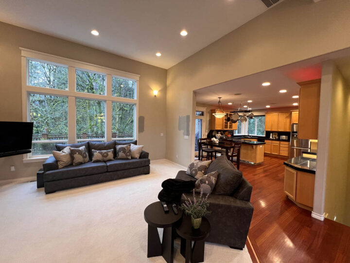 Spacious living room with a large gray sectional sofa, hardwood floors, and adjacent open kitchen with warm lighting. large windows offer a view of greenery outside.