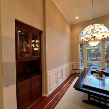 Dining room with wooden flooring and a large window, featuring a chandelier, wooden cabinet, and a glass-topped table.