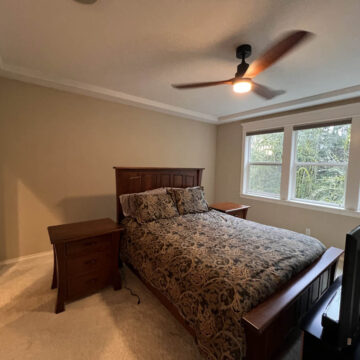 A bedroom featuring a wooden bed with patterned bedding, a nightstand, a ceiling fan, and large windows showing greenery outside.