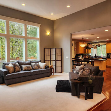 Modern living room with large windows overlooking trees, connected to a kitchen with wooden cabinets and black countertops.