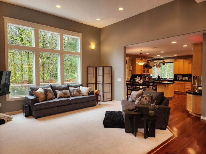 Modern living room with large windows overlooking trees, connected to a kitchen with wooden cabinets and black countertops.