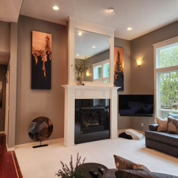 A modern living room featuring a fireplace, mounted tv, and comfortable seating from an interior painting project in Tigard that brightened the space, with large windows overlooking greenery outside.