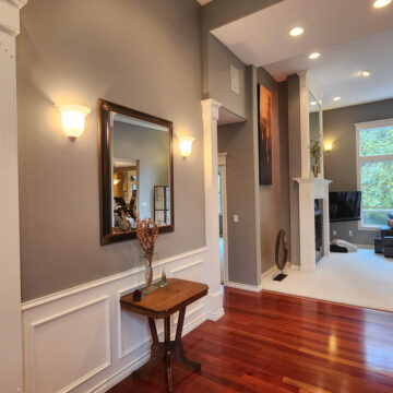 Interior of a house showing a hallway with hardwood floors, gray walls, white trim, wall-mounted lamps, a mirror, and an open door leading to a living room.