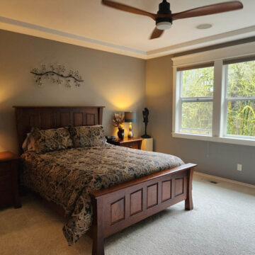 A cozy bedroom with a large bed, wooden furniture, and a ceiling fan, overlooking a window with leafy views.