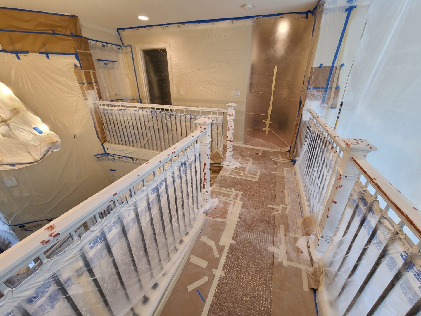 A staircase area under renovation, covered with protective plastic sheets and tape, showcases partially painted railings and walls. Discover how to spray for professional results as you navigate spray trim techniques that ensure a flawlessly finished look.