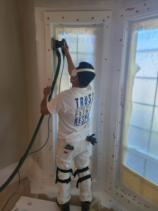 Person with protective earmuffs sanding a window frame inside a room, wearing a white t-shirt and pants, with a vacuum sander attached.