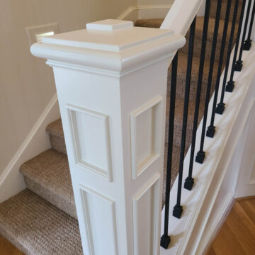 White staircase with a decorative post, black metal balusters, and carpeted steps.