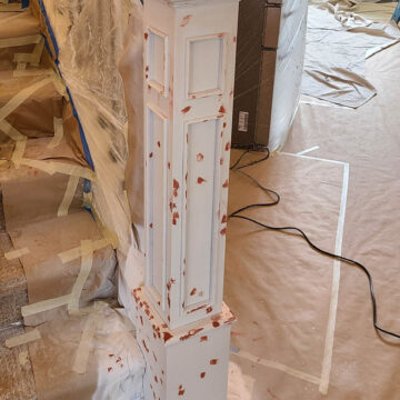 A white wooden newel post with areas of red filler, surrounded by drop cloths and taped surfaces, indicating ongoing renovation work.