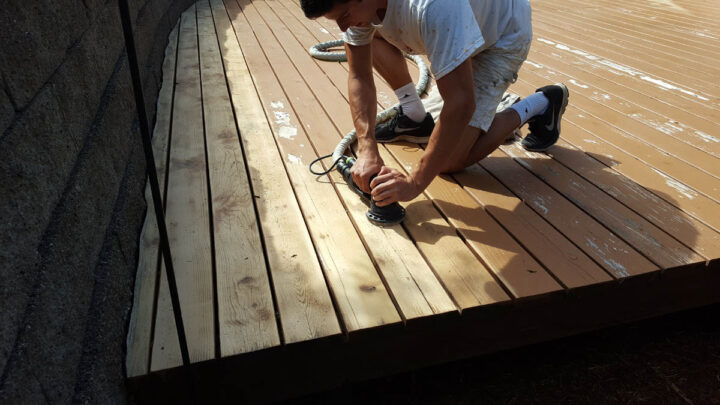 A person kneels on a wooden deck, using a power sander to smooth the surface. The deck appears partially sanded, with a visible contrast between sanded and unsanded areas.