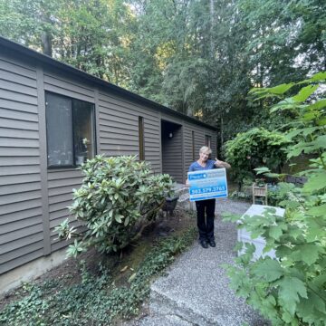 A woman standing outside of a Portland home with a sign promoting their Exterior Paint Project.