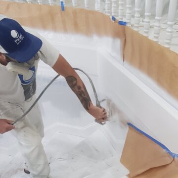 A person spray painting a white staircase, donning a cap, mask, and protective clothing, with taped paper shielding nearby cabinets.