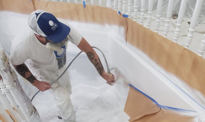 A person spray painting a white staircase, donning a cap, mask, and protective clothing, with taped paper shielding nearby cabinets.