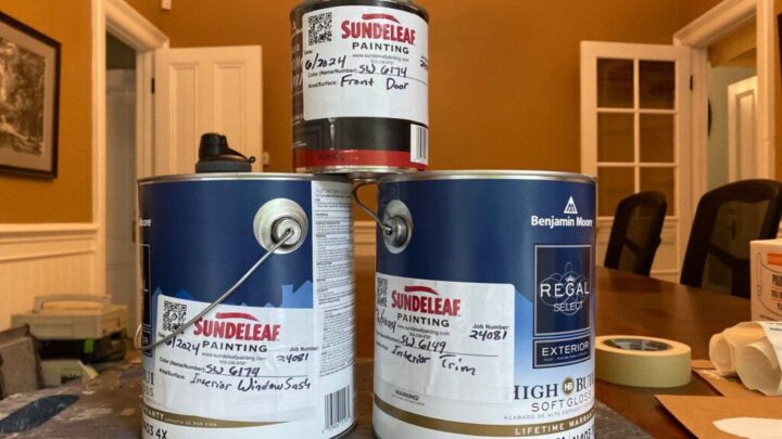 In a room with yellow walls and wooden chairs, Portland painters have neatly stacked paint cans, each labeled with details on type and usage for a front door and exterior trim.