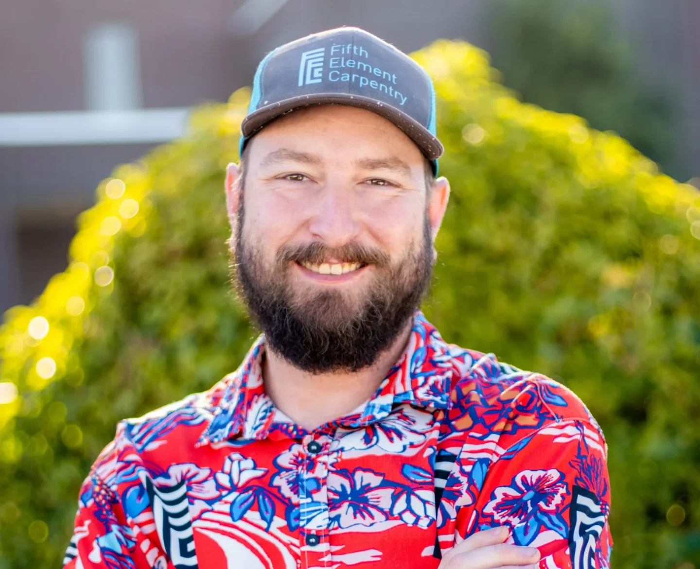 A bearded man in a "Fifth Element Carpentry" hat and red floral shirt stands outside in Portland, smiling with arms crossed.